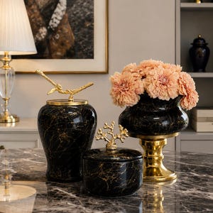 May include: Two black marble-patterned ceramic jars with gold-toned lids featuring branch and coral-like finials, alongside a black marble-patterned bowl on a gold pedestal base filled with a cluster of peach-colored artificial flowers.