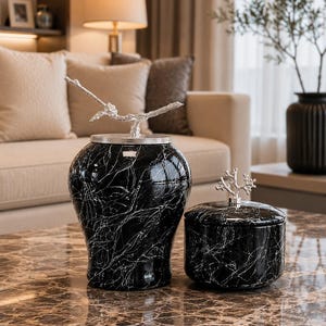 Può includere: Due barattoli decorativi neri con motivo marmorizzato e coperchi argentati. Il barattolo più alto presenta una maniglia argentata a forma di ramo, mentre quello più basso e largo ha una maniglia a forma di corallo. Finitura lucida.