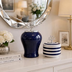 May include: A glossy dark blue ceramic jar with a silver branch-shaped lid handle, positioned next to a white and dark blue striped ceramic container featuring a silver tree-shaped lid handle, both resting on a smooth white surface.