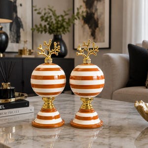 Puede incluir: Dos objetos decorativos con cuerpo esférico de rayas horizontales naranjas y blancas, apoyados sobre una base de pedestal dorada y rematados con un adorno dorado en forma de coral, con un acabado liso y brillante.