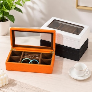 May include: A rectangular jewelry box with an orange exterior and a hinged lid featuring a clear glass window. The interior is lined with brown fabric and divided into multiple compartments, including a section for rings and two blue bracelets.