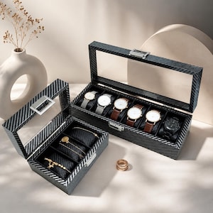 May include: Two black carbon fibre patterned storage boxes with clear glass lids and silver metal latches. The larger box holds six wristwatches with various straps, while the smaller box contains three black velvet cushions displaying gold jewellery.