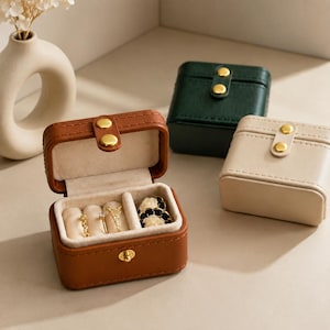 May include: Three small square jewelry boxes in brown, dark green, and cream. Each box features a gold-tone snap closure and a soft beige interior lining. The open brown box displays three gold rings and a pair of floral-shaped earrings inside.
