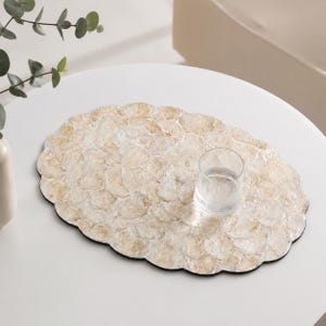 May include: An oval placemat constructed from overlapping, translucent, light beige and white shell-like discs with a scalloped edge. A clear, cylindrical glass tumbler sits on the right side of the textured, layered surface of the placemat.