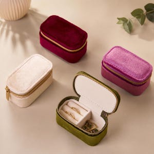 May include: Four rectangular velvet jewellery cases with gold-tone zips. Three cases are closed in deep red, cream, and magenta, while one olive green case is open, revealing a cream-coloured interior with ring slots and a small storage compartment.