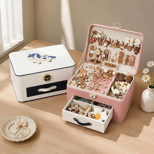 May include: A white jewelry box with a blue unicorn graphic and a pink jewelry box with an open lid and drawer, both featuring gold-tone latches. The pink box contains various hair accessories, clips, and bands in brown, white, and cream tones.