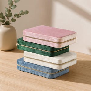 May include: A stack of four rectangular jewelry cases covered in soft velvet fabric. The cases are colored in shades of dusty pink, dark green, cream, and dusty blue. Each case features a gold-toned zipper closure running along the side perimeter.