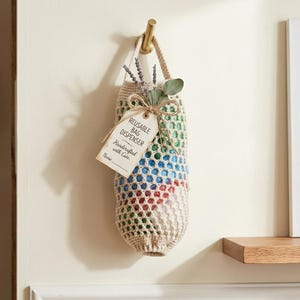 Peut inclure: Un distributeur de sacs réutilisables en crochet beige suspendu à un crochet doré. Le support cylindrique présente un motif en maille contenant des sacs en plastique colorés. Une étiquette en papier est attachée avec de la ficelle, REUSABLE BAG DISPENSER.