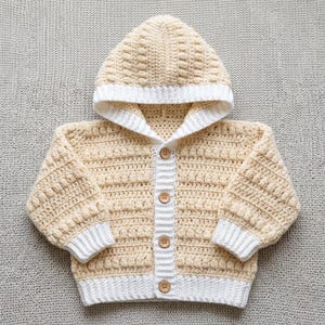 May include: A cream-coloured crochet hooded cardigan featuring a textured bobble stitch pattern throughout the body and sleeves. The garment includes a white ribbed trim along the hood, cuffs, and hem, and is fastened with four light brown wooden buttons.