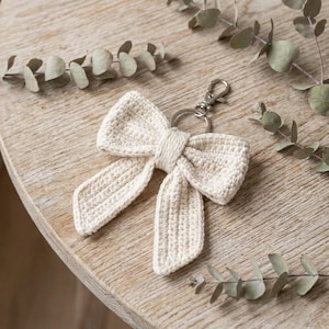 May include: A cream-colored crochet bow keychain featuring a textured, hand-stitched yarn construction. The bow is attached to a silver-toned metal split ring and a lobster claw clasp, providing a secure attachment point for keys or accessories.