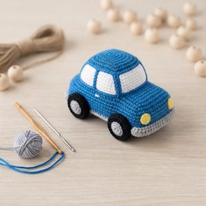 May include: A blue crocheted toy car with white windows, black and gray wheels, and yellow headlights. The car is crafted with visible yarn stitches, featuring a rounded body shape and a gray bumper, showcasing a detailed handmade textile design.