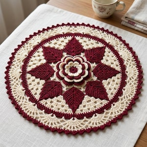 Peut inclure: Un napperon en crochet rond présentant une fleur centrale superposée en crème et rouge foncé, entourée d'un anneau de huit motifs en forme de feuilles rouge foncé. La bordure extérieure se compose de dentelle ajourée crème à bord festonné rouge.