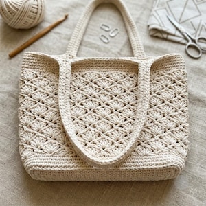Peut inclure: Un sac cabas en crochet de couleur crème présentant un motif répétitif de points en éventail sur tout le corps, une base solide et deux anses robustes. Le sac est fabriqué à partir de fil épais et affiche un design texturé et ajouré.