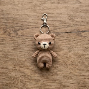 May include: A light brown crocheted teddy bear keychain featuring a small cream-colored muzzle, black embroidered nose and mouth, and two black bead eyes. The bear is attached to a silver-toned metal split ring and a lobster claw clasp fastener.