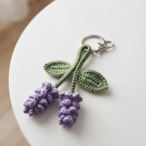 May include: A crochet keychain featuring two lavender-colored flower clusters attached to green stems with two green leaves. The stems are joined at the top to a silver-toned metal split ring, providing a functional attachment point for keys.