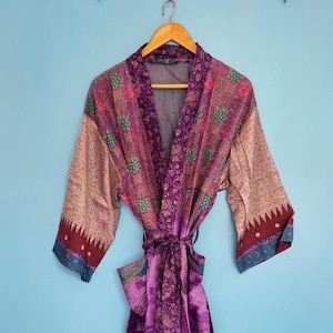 Könnte beinhalten: Ein langärmeliger Morgenmantel mit einem lebendigen Patchwork-Design in Lila-, Rosa- und Türkistönen. Das Kleidungsstück verfügt über einen passenden Bindegürtel in der Taille und einen breiten Kragen mit einer Mischung aus floralen Mustern.