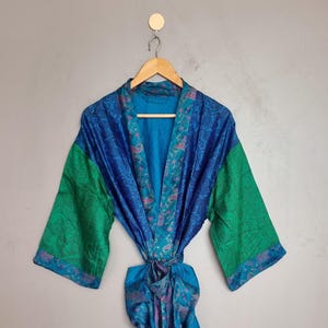 May include: A long-sleeved robe featuring a vibrant blue and green color palette with intricate paisley patterns. The garment includes a matching tie belt at the waist, wide sleeves, and a shawl collar, all displayed on a wooden hanger.