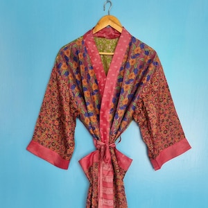 Könnte beinhalten: Ein langärmeliger Kimono-Bademantel mit Patchwork-Design und Blumenmustern in Rosa-, Orange-, Lila- und Grüntönen. Das Kleidungsstück verfügt über einen einfarbigen rosa Kragen und einen passenden Bindegürtel und hängt an einem Holzkleiderbügel.