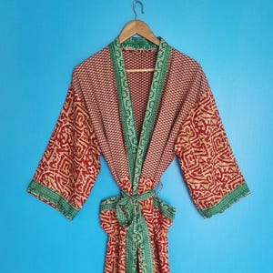Könnte beinhalten: Ein langärmeliger Morgenmantel aus einem rot-beigen Stoff mit einem kontrastierenden grün-weißen Blumenmuster entlang der Vorderseite, der Ärmelbündchen und des Bindegürtels. Das Kleidungsstück hängt auf einem Holzkleiderbügel vor blauem Grund.