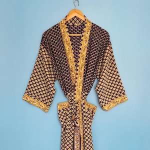Op de afbeelding: Een kamerjas met lange mouwen en een donkerpaarse basis met een herhalend goudkleurig bladvormig patroon. Het kledingstuk heeft een contrasterende gouden rand met een bloemenprint langs de opening aan de voorzijde, de mouwen en een ceintuur.