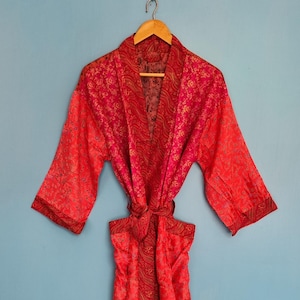 Puede incluir: Una bata de manga larga en tonos vibrantes de rojo y rosa, con un intrincado patrón floral y de cachemira repetitivo. La prenda incluye un cinturón de tela a juego en la cintura y un cuello de solapa ancho y contrastante.