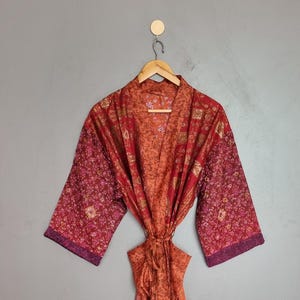 May include: A long-sleeved robe featuring a patchwork design with various shades of red, orange, and purple. The garment includes a tie-waist belt and a floral pattern throughout the fabric, hanging from a wooden hanger against a grey wall.