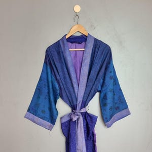 May include: A long-sleeved robe in shades of blue and purple, featuring a wrap-around front with a matching tie belt. The garment displays a subtle, repeating geometric pattern throughout the fabric, with wide sleeves and a structured collar.