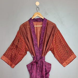 May include: A long-sleeved robe featuring a mix of deep purple and burnt orange fabric. The garment includes a purple paisley-patterned collar and matching waist tie, with sleeves displaying a contrasting orange and red geometric print.