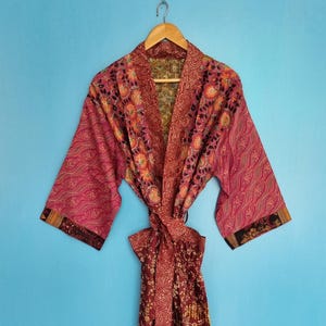 Op de afbeelding: Een kamerjas met lange mouwen voorzien van een patchwork van bloem- en geometrische patronen in tinten dieprood, roze en goud. Het kledingstuk heeft een brede kraag, een bijpassende ceintuur in de taille en contrasterende donkere mouwboorden.