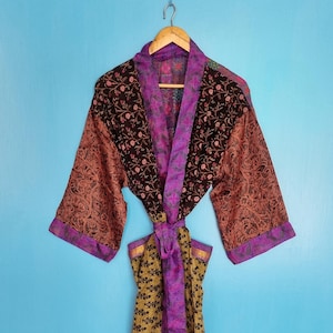 Peut inclure: Une robe à manches longues présentant un motif patchwork avec une partie supérieure florale marron foncé, des manches à motifs cuivrés et une partie inférieure florale jaune moutarde. Le vêtement inclut une bordure violet vif et une ceinture.