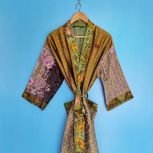 Könnte beinhalten: Ein langärmeliger Morgenmantel mit Patchwork-Design, bestehend aus verschiedenen Blumen- und geometrischen Mustern in Olivgrün, Braun, Lila und Gelb. Das Kleidungsstück verfügt über einen passenden Bindegürtel in der Taille und einen breiten Kragen, hängend an einem Holzkleiderbügel.