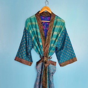 Könnte beinhalten: Ein langärmeliger Kimono-Bademantel aus einem Patchwork aus blaugrünen, blauen und violetten Stoffen mit komplizierten Mustern. Das Kleidungsstück hat einen dunkelbraunen Kragen mit Muster und einen passenden Bindegürtel, hängend an einem Holzkleiderbügel vor einer blauen Wand.