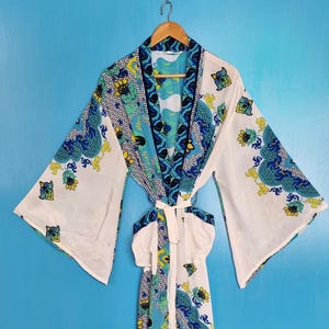 Könnte beinhalten: Ein langärmeliger Morgenmantel mit weißer Grundfarbe und einem lebhaften blauen, gelben und grünen Blumen- und Abstraktmuster. Das Kleidungsstück hat einen breiten Kragen, weite Ärmel und einen passenden Bindegürtel in der Taille, hängend an einem Holzkleiderbügel.