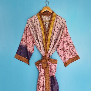 Könnte beinhalten: Ein knielanger Kimono-Bademantel mit einem Patchwork-Design aus rosa, violetten und braunen Blumenmustern. Das Kleidungsstück verfügt über weite Ärmel, eine braun gemusterte Borte entlang der Vorderseite und einen passenden Bindegürtel.