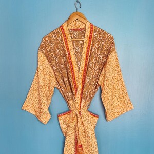 May include: A long-sleeved robe featuring a mix of patterns in shades of orange, brown, and cream. The garment has a wrap-front design with a tie belt at the waist, a shawl collar, and contrasting orange and red trim along the front edges.