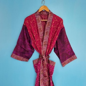 Könnte beinhalten: Ein langärmeliger Morgenmantel in leuchtendem Magenta und tiefem Violett mit komplizierten Paisley- und Blumenmustern. Das Kleidungsstück verfügt über einen breiten Schalkragen, einen passenden Bindegürtel in der Taille und weite Ärmel aus leichtem Stoff.