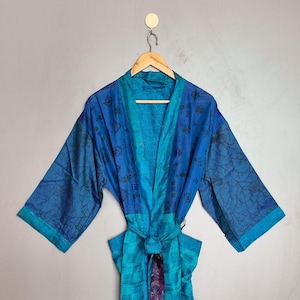 May include: A long-sleeved kimono robe in shades of blue and teal, featuring a patchwork design with varied patterns including geometric squares, floral motifs, and branch-like prints. The garment includes a matching tie belt and a dark purple trim.