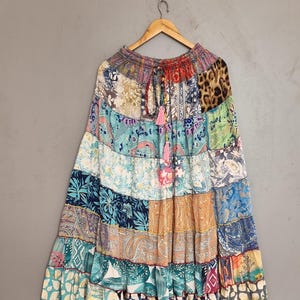 May include: A long, tiered patchwork skirt featuring a variety of colorful fabric patterns, including floral, paisley, and animal prints. The skirt has an elasticized waistband with a drawstring and two pink tassels hanging from the center front.