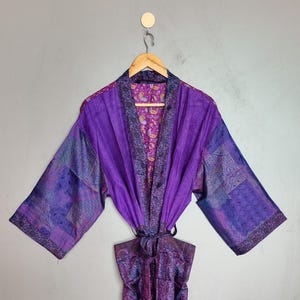 Könnte beinhalten: Ein langärmeliger Morgenmantel in verschiedenen Violett- und Purpurtönen mit einem Patchwork-Design aus diversen komplexen Mustern. Das Kleidungsstück verfügt über einen breiten Kragen, einen Bindegürtel und ein sichtbares Innenfutter mit Blumenmuster.