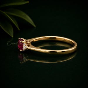 May include: A gold ring featuring a central oval-cut red gemstone accented by a cluster of small clear stones on one side. The band is smooth and polished, showcasing a reflective surface and a delicate setting that holds the stones securely.