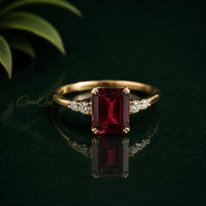 May include: A gold ring featuring a central emerald-cut deep red gemstone flanked by two small round white stones on each side. The band is polished gold, and the text Orin Oak is visible on the left side of the ring setting.
