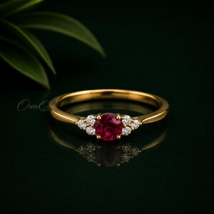 Puede incluir: Un anillo de oro con una piedra central de rubí rojo intenso de forma redonda, flanqueada por tres pequeños diamantes redondos y transparentes a cada lado. La banda es de oro amarillo pulido y el engaste sostiene las piedras en un grupo.