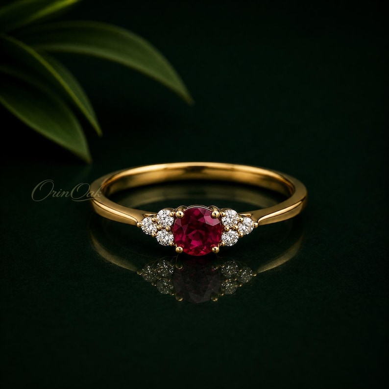 May include: A gold ring featuring a round, deep red ruby center stone flanked by three small, clear, round-cut diamonds on each side. The band is polished yellow gold, and the setting holds the stones in a cluster arrangement on a dark background.