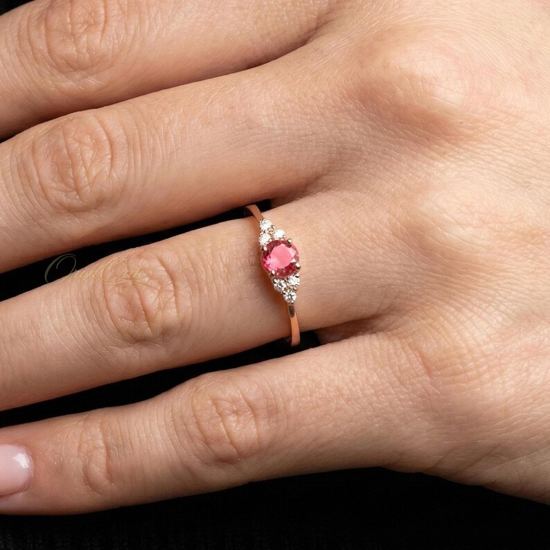 May include: A rose gold ring featuring a central round-cut pink gemstone flanked by three small clear stones on each side. The band is thin and polished, showcasing a delicate design that highlights the vibrant pink center stone and the surrounding clear accents.