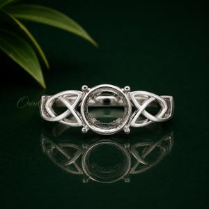 Könnte beinhalten: Eine silberfarbene Ringfassung mit einer zentralen runden Halterung für einen Edelstein, die auf beiden Seiten von komplizierten, durchbrochenen keltischen Knotenmustern flankiert wird. Das Metall hat eine polierte und reflektierende Oberfläche.