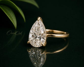 Könnte beinhalten: Ein Ring mit einem birnenförmigen Diamanten in einer polierten Gelbgoldschiene. Der große, facettierte Stein wird von drei Krappen gehalten und reflektiert das Licht. Der Ring ist vor einem dunklen Hintergrund mit dem Orin Oak Logo platziert.