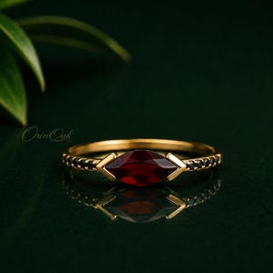 Op de afbeelding: Een gouden ring met een centrale dieprode granaat edelsteen in marquise-slijpvorm, vastgehouden door twee chevronvormige zettingen, geflankeerd door een rij kleine zwarte steentjes langs de dunne ringband tegen een donkergroene achtergrond.