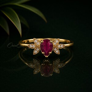 May include: A gold ring featuring a central pear-shaped deep red ruby gemstone flanked by two leaf-shaped accents set with small round white diamonds. The band is polished gold, and the design includes the text Orin Oak on the left side.