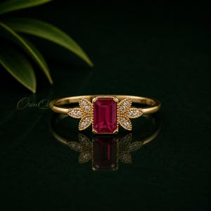 May include: A gold ring featuring a central emerald-cut red gemstone flanked by two leaf-shaped accents, each set with small round white stones. The band is thin and polished gold, with the text Orin Oak visible on the left side of the ring.