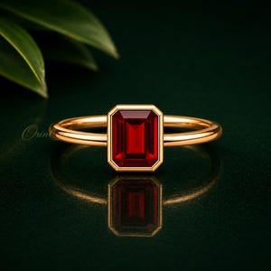 May include: A gold ring featuring a single emerald-cut deep red gemstone set in a polished gold band. The ring is centred horizontally, showcasing the rectangular stone's facets and the smooth, rounded profile of the metal band against a dark surface.